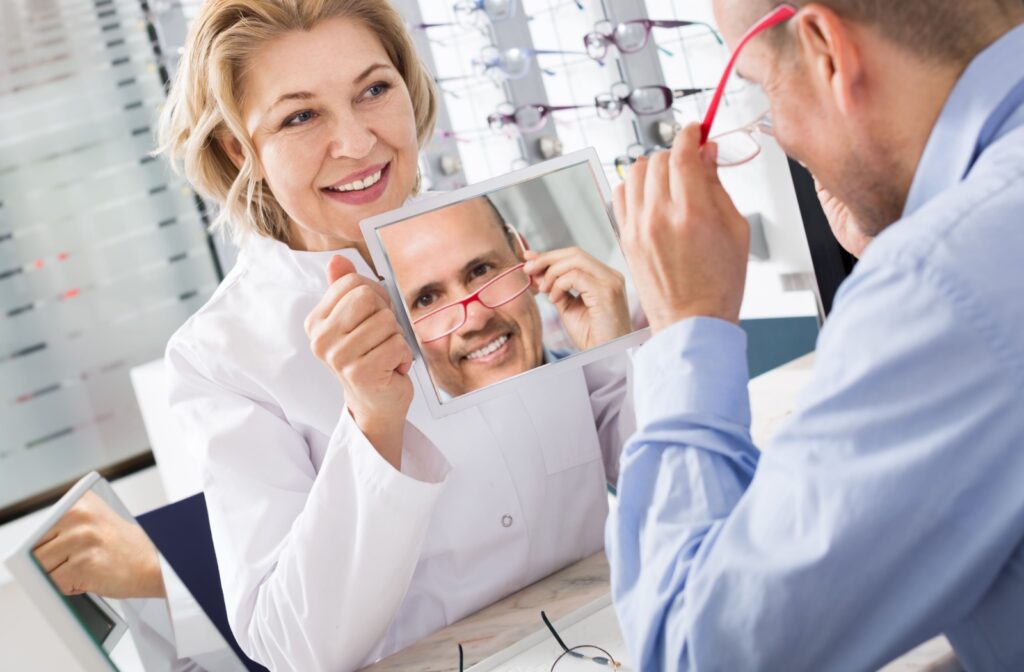 An optometrist helps a man try on red eyeglass frames using a handheld mirror.
