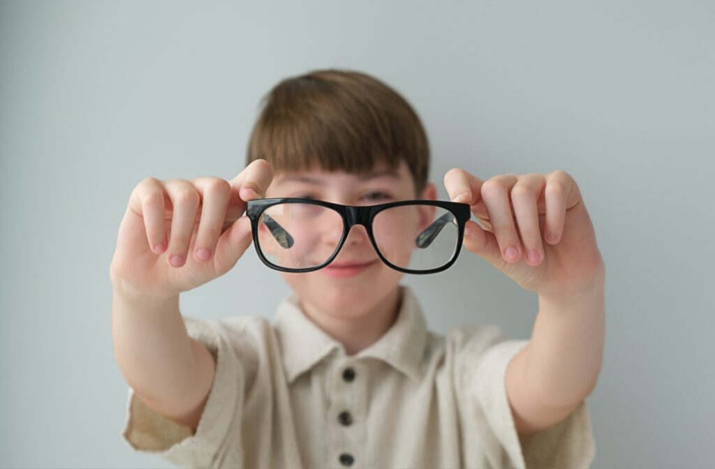 A young child holding up a pair of black eyeglasses in front of their face, smiling slightly, with a light gray background.